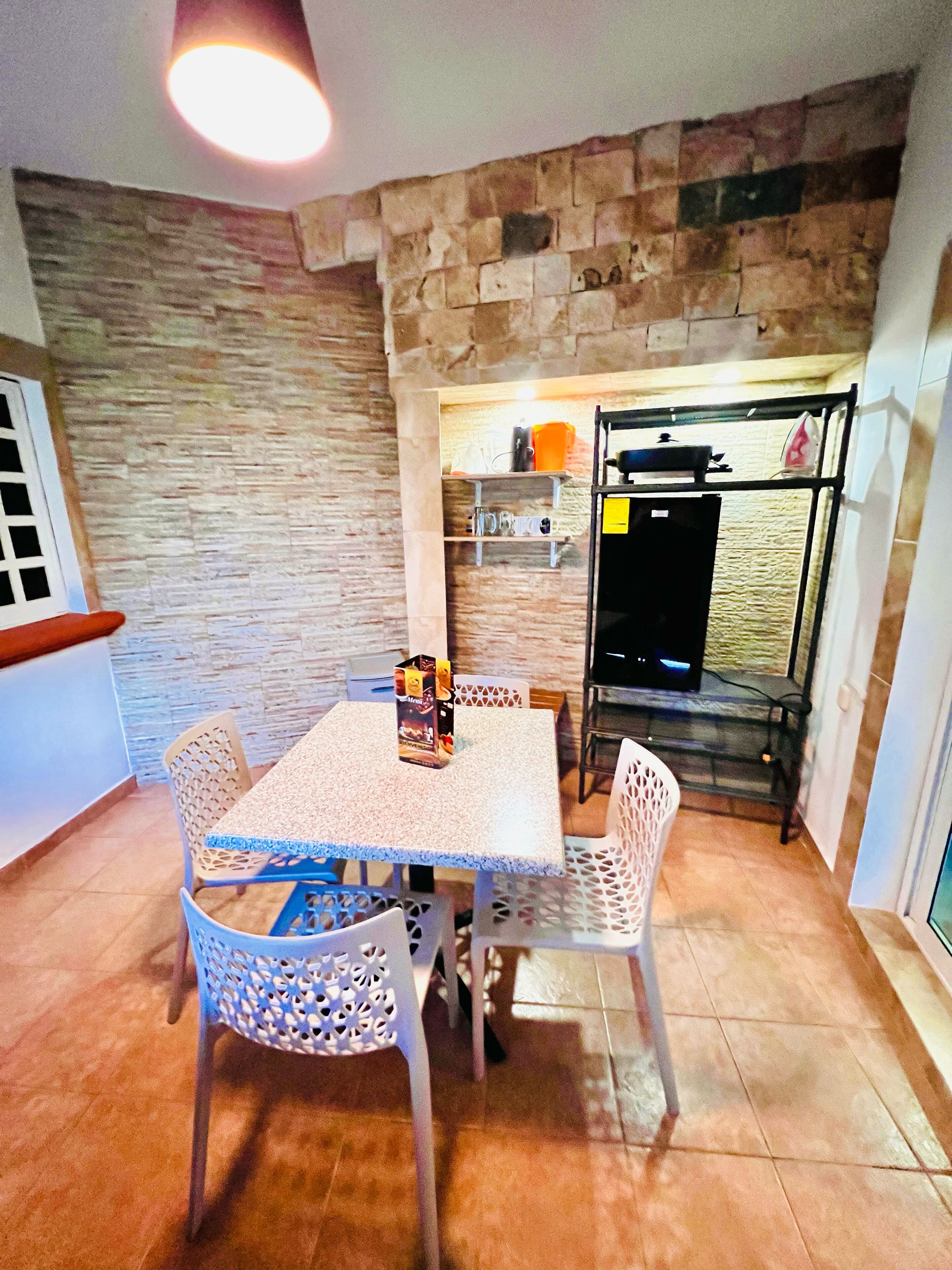 Un comedor con una mesa y sillas, una pared de piedra y una zona de cocina con refrigerador y estantes.