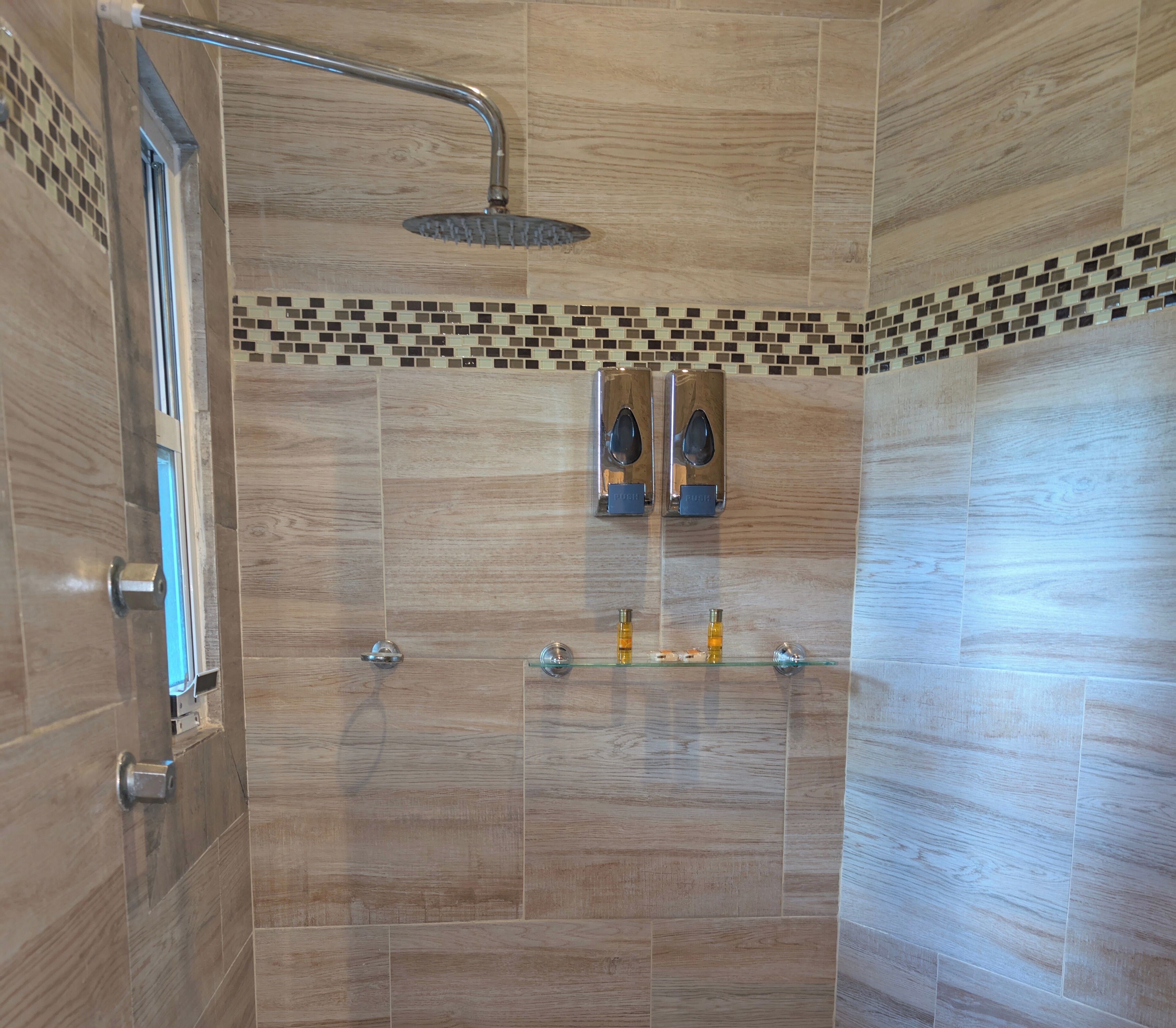 Una doccia moderna con soffione a pioggia, uno scaffale in vetro con articoli da toilette e una parete piastrellata con bordo a mosaico.