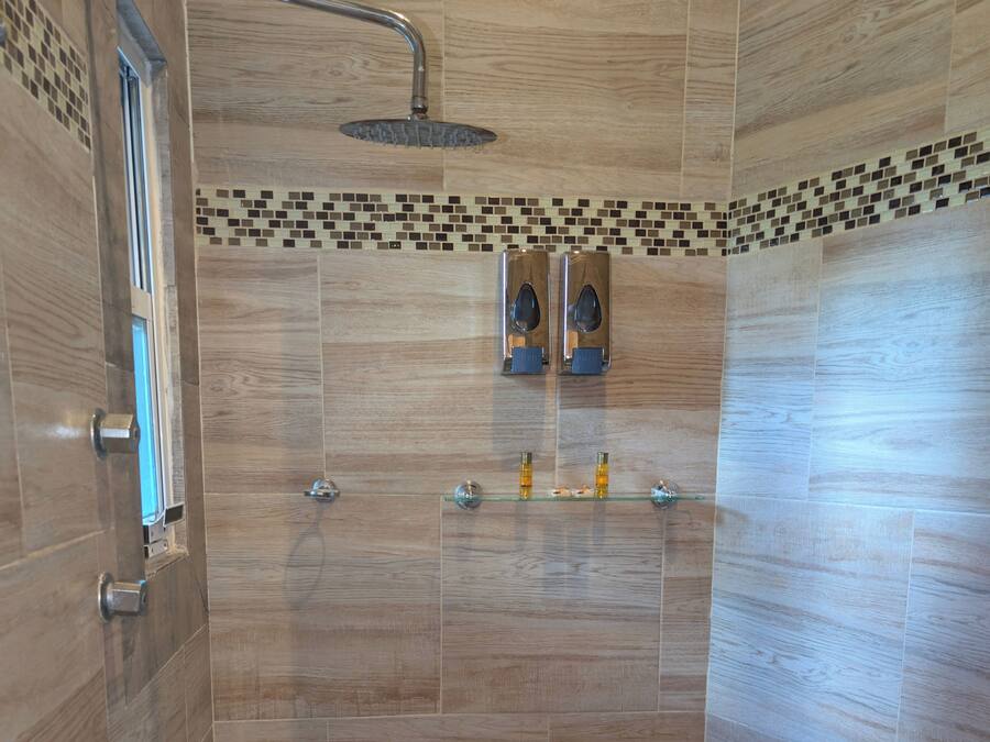 Una doccia moderna con soffione a pioggia, uno scaffale in vetro con articoli da toilette e una parete piastrellata con bordo a mosaico.