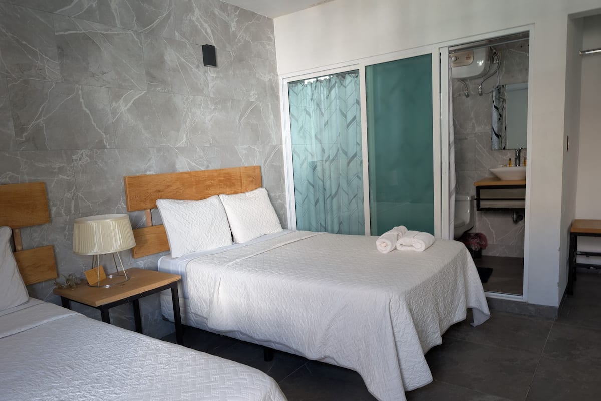 A hotel room with two beds, a wooden headboard, a nightstand with a lamp, a bathroom with a sink and mirror, and a sliding glass door to the shower area.