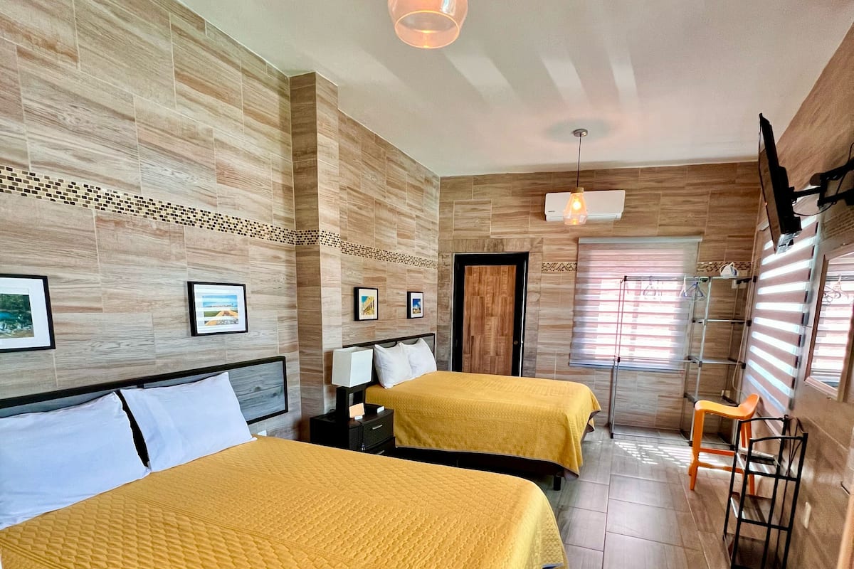 A hotel room with two beds, wooden flooring, a hanging light fixture, and a window with blinds.