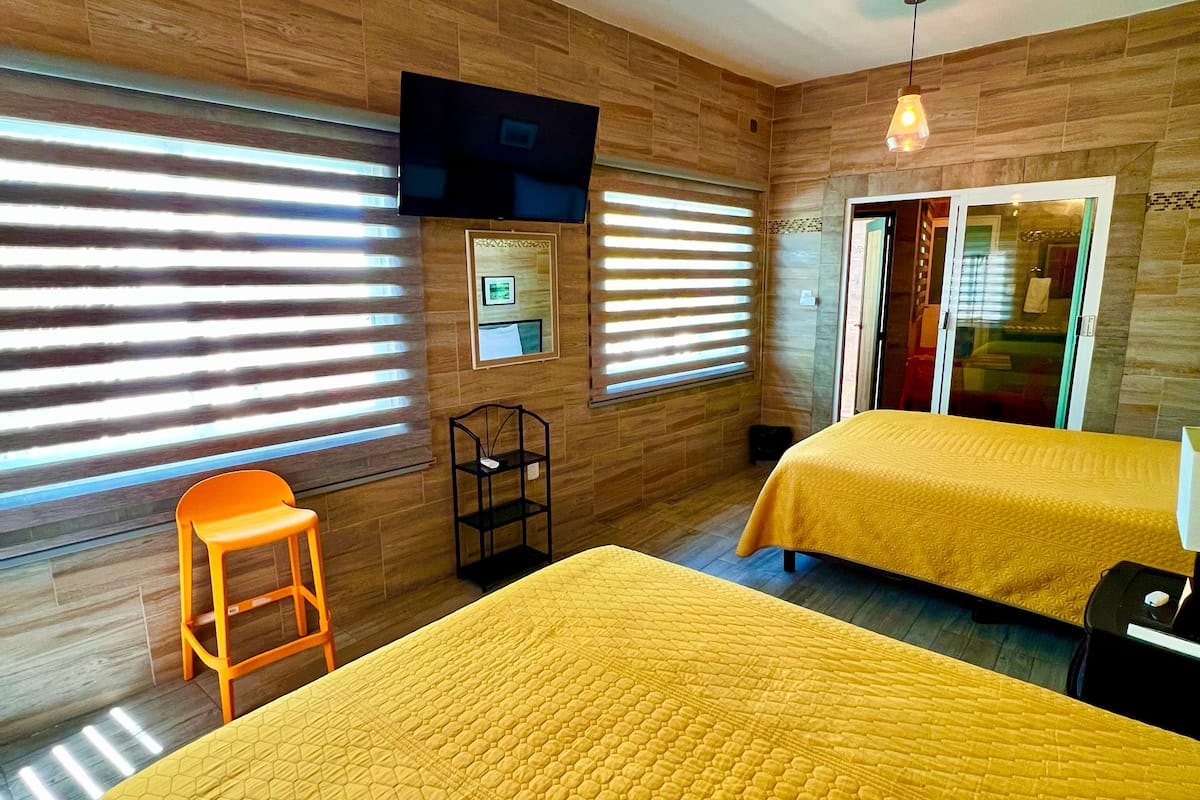 A hotel room with wooden walls, two beds with yellow bedding, a TV mounted on the wall, and a window with blinds.
