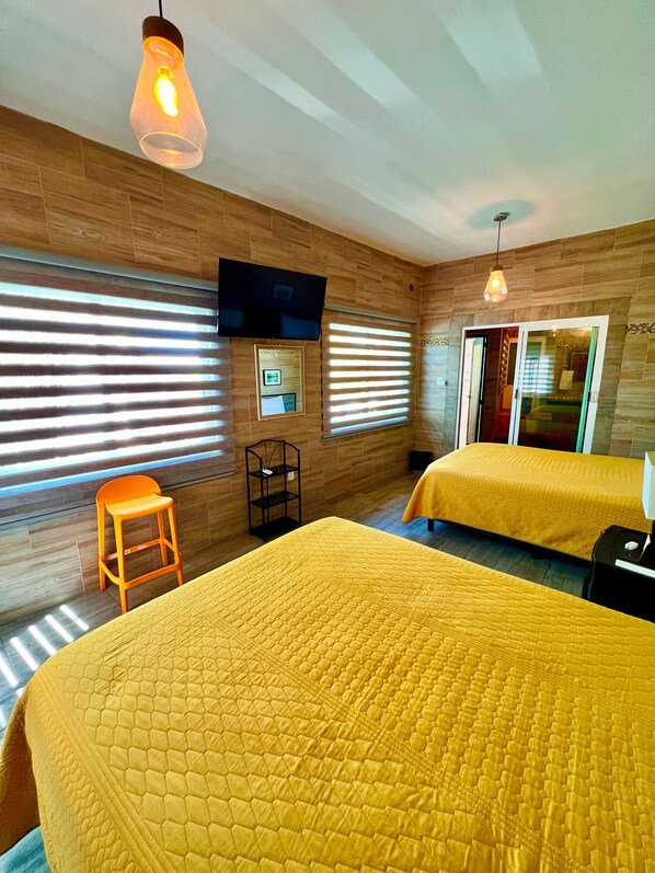 A hotel room with wooden walls, two beds with yellow bedding, a TV mounted on the wall, and a window with blinds.
