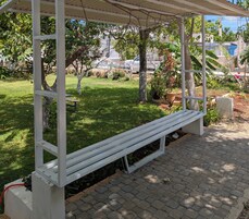 Un espacio de estar al aire libre cubierto con un banco, rodeado de un jardín con diversas plantas y árboles.