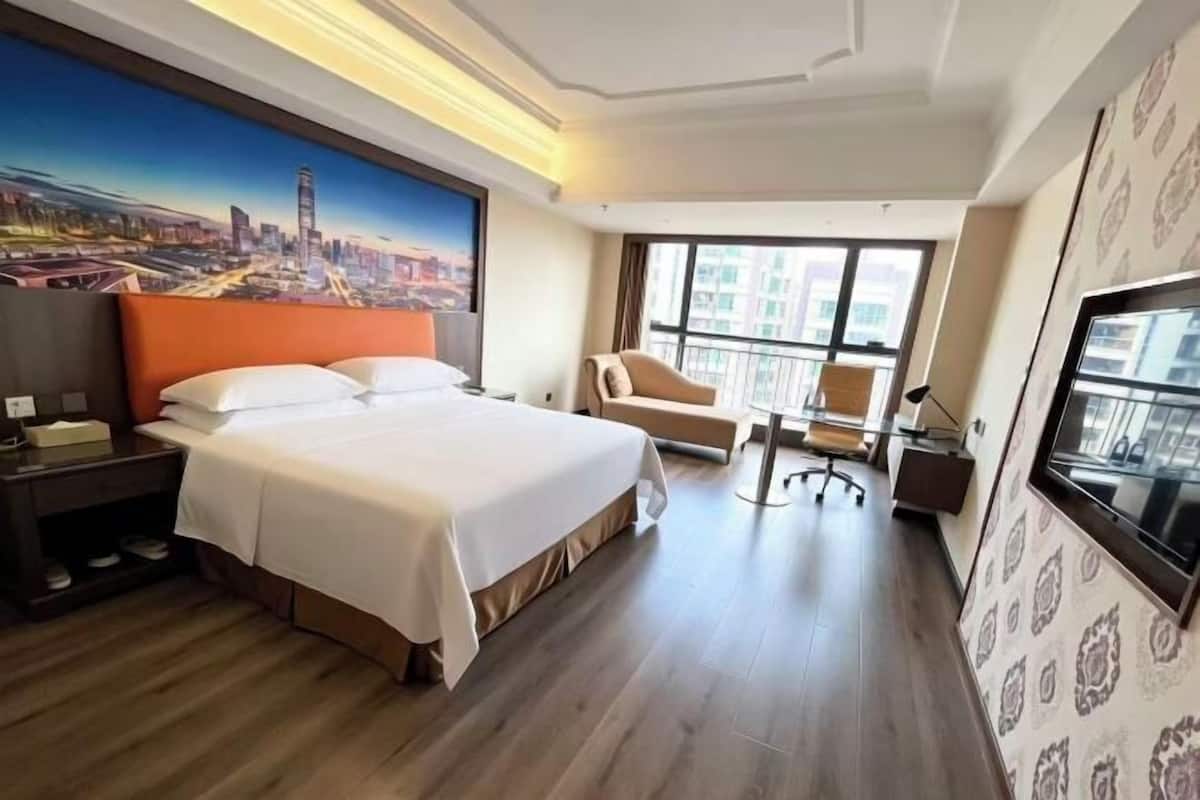 A hotel room with a large bed, a chair, a desk, a TV, and a cityscape painting on the wall.
