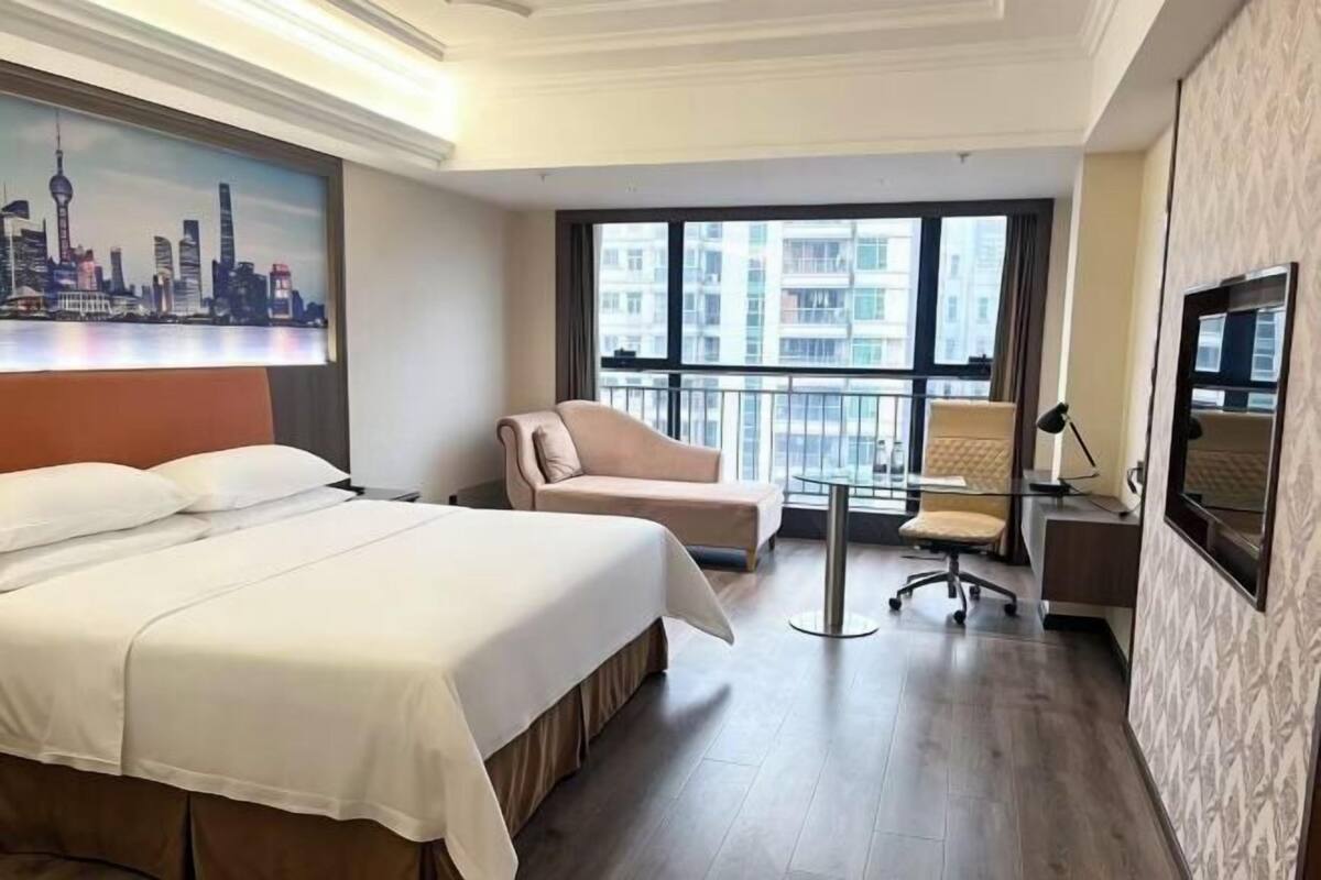 A hotel room with a bed, a sofa, a desk, and a chair. There is a large window with a view of buildings.