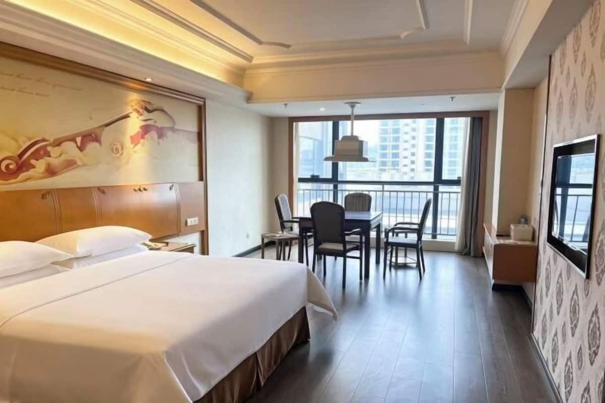 A hotel room with a large bed, a desk with chairs, a TV, and a balcony with a city view.