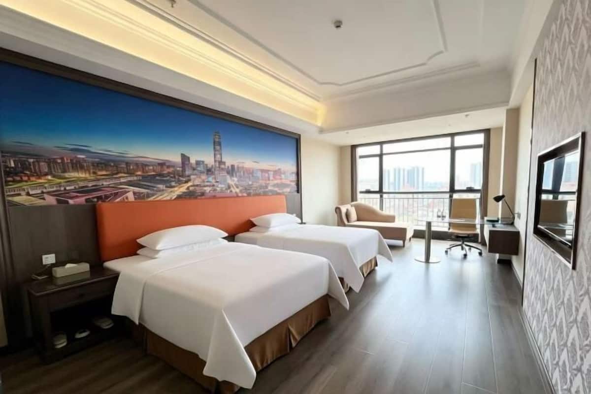 A hotel room with two beds, a desk, a chair, a large window, and a cityscape painting on the wall.
