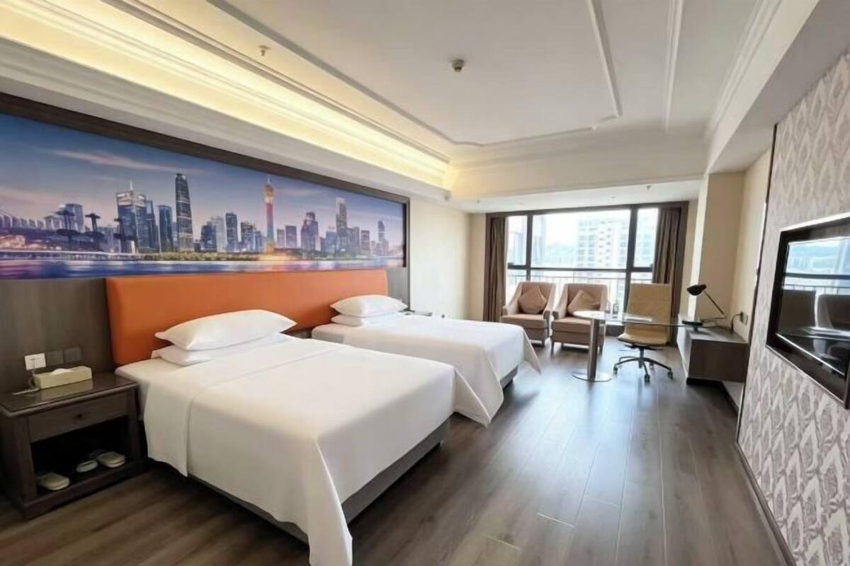 A hotel room with two beds, a desk, a chair, a TV, and a cityscape painting on the wall.