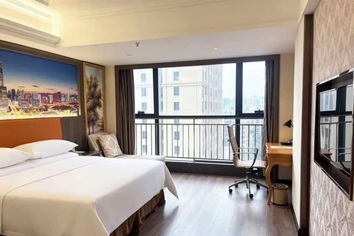 A hotel room with a large bed, a desk, a chair, a window with a city view, and a painting on the wall.