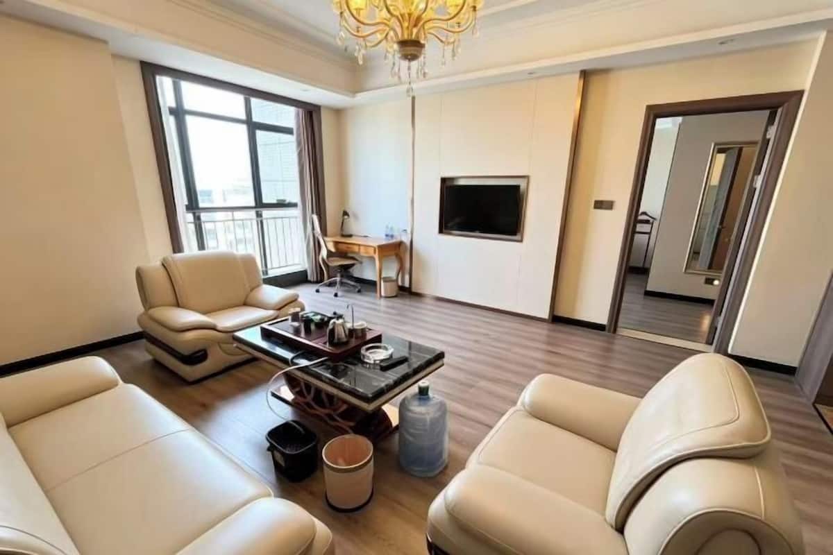A spacious living room with leather sofas, a coffee table, a TV mounted on the wall, and a large window providing natural light.