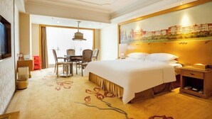 A hotel room with a large bed, a desk with chairs, a dining area, and a painting on the wall. - Vienna International Hotel (Zhongshan Torch Development Zone (Zhongshan)