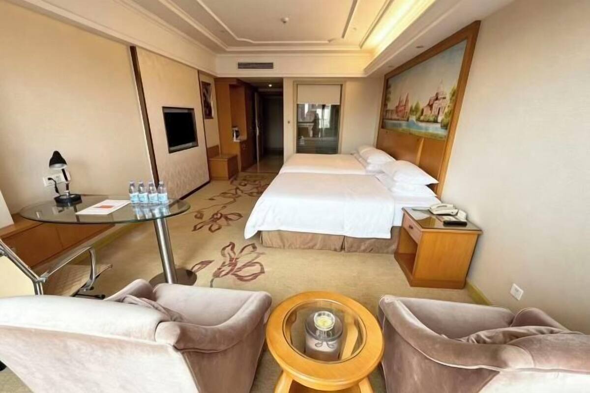 A hotel room with a bed, a desk with a lamp, a chair, a small table, and a painting on the wall.