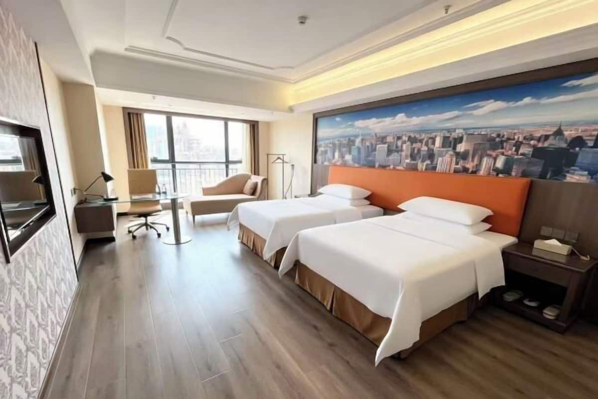 A hotel room with two beds, a desk, a chair, a TV, and a large cityscape painting on the wall.