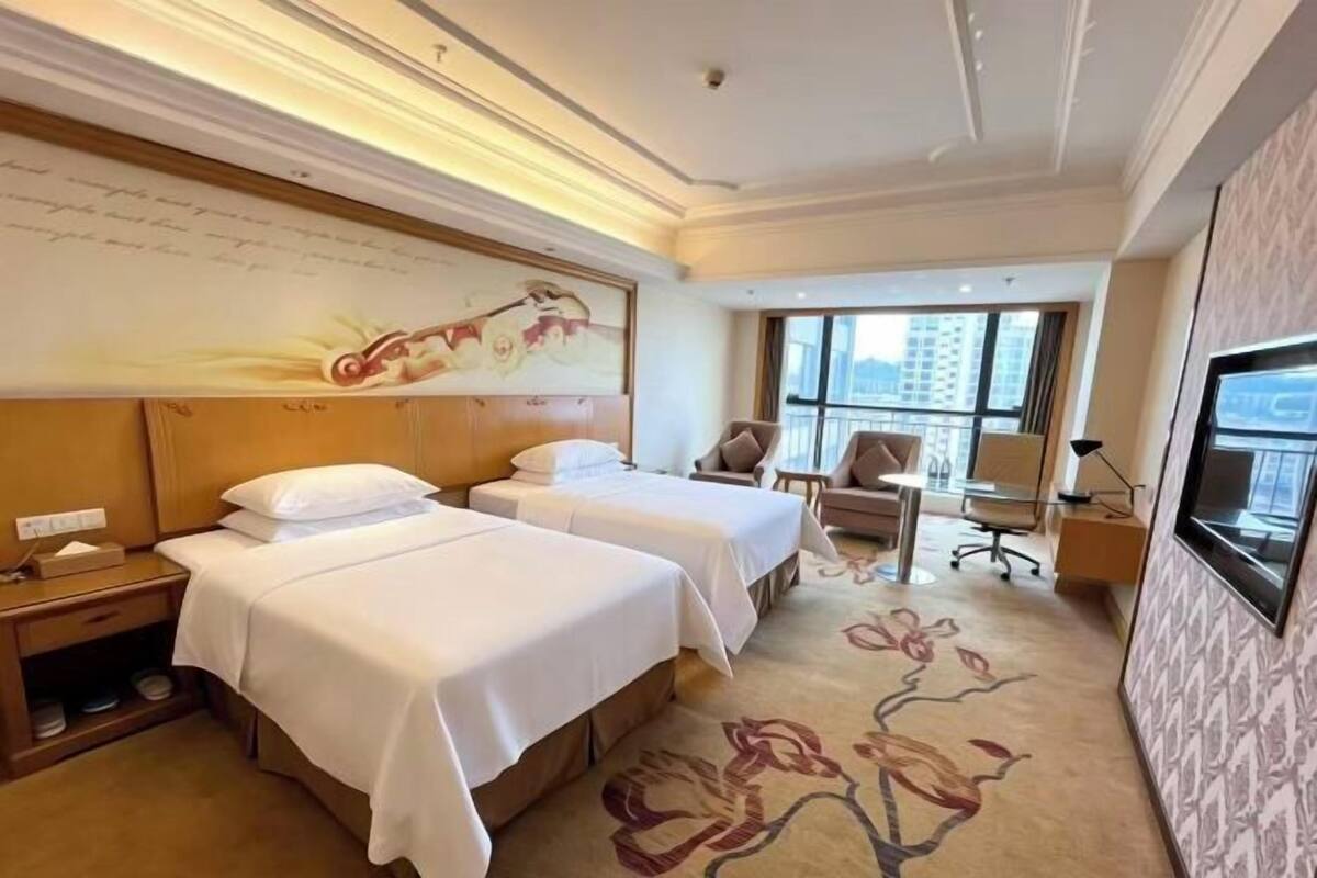 A hotel room with two beds, a desk, a chair, a TV, and a large window.