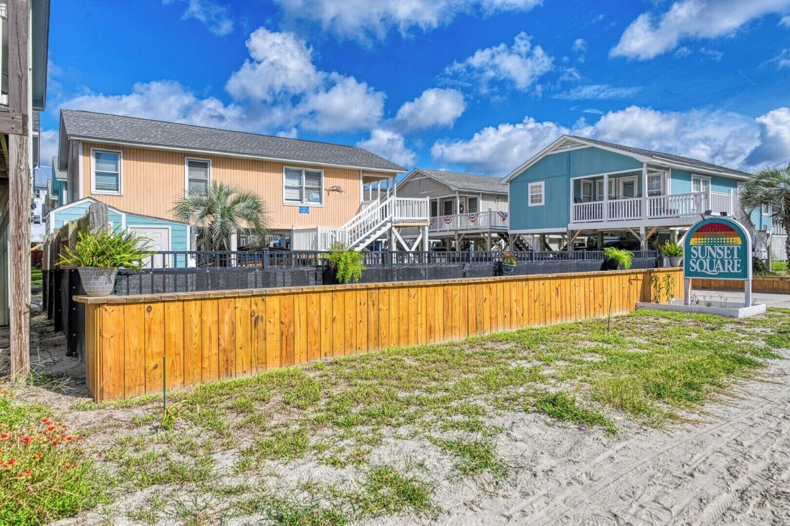 A beachfront property with a wooden fence, a sign reading "Sunset Square," and a building with a balcony.