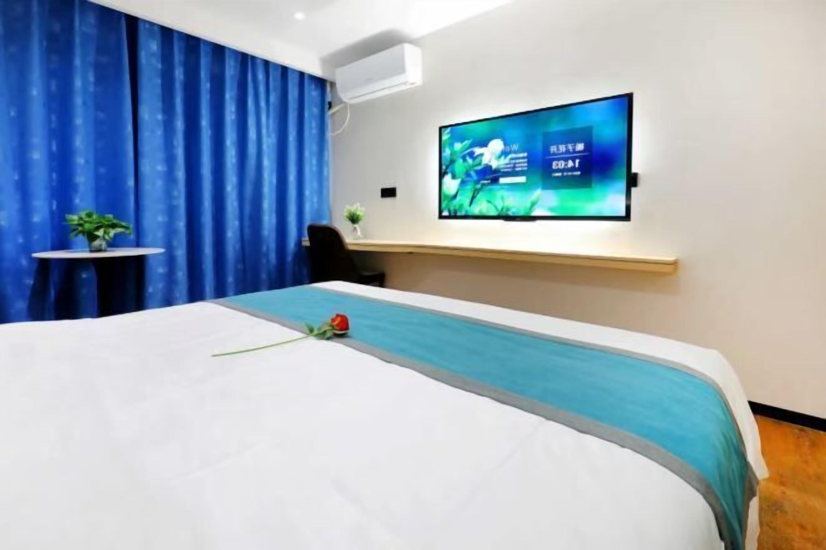 Quarto de hotel com cama, televisão na parede, mesa pequena com uma planta e escrivaninha com cadeira.