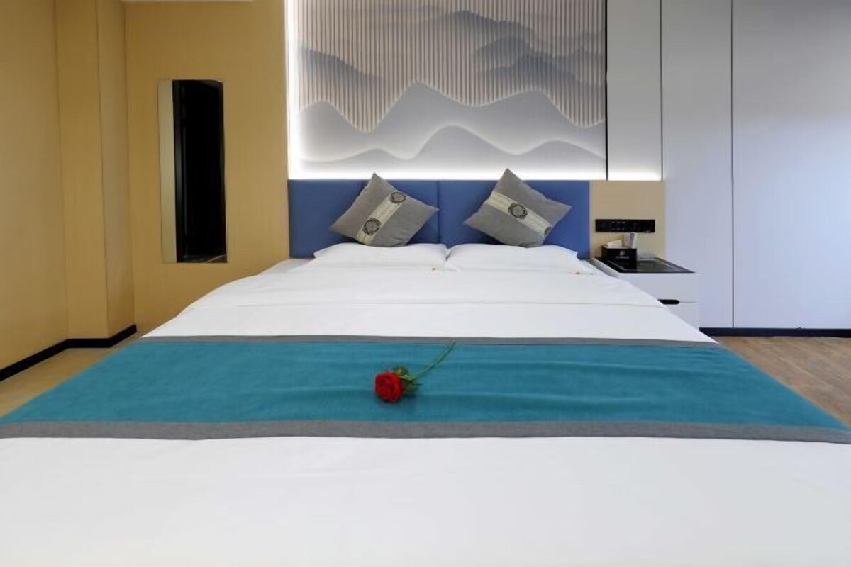 A neatly made bed with a blue accent runner, a single red rose, and a wall-mounted light fixture.