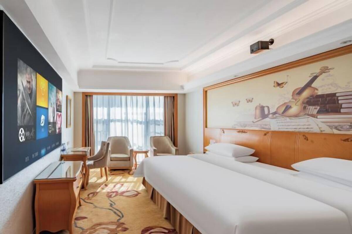 A hotel room with a large bed, a desk, two chairs, a TV, and a mural on the wall.