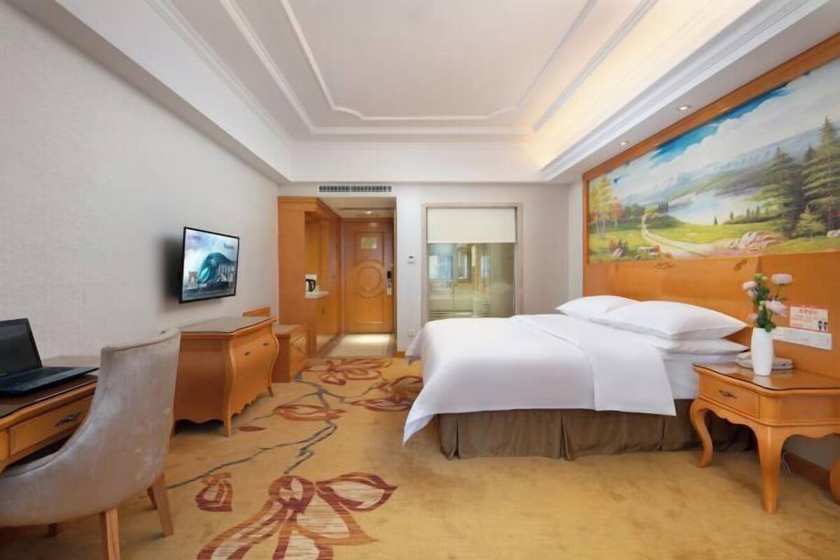 A hotel room with a bed, a desk with a laptop, a TV, a painting on the wall, and a carpet with a floral pattern.
