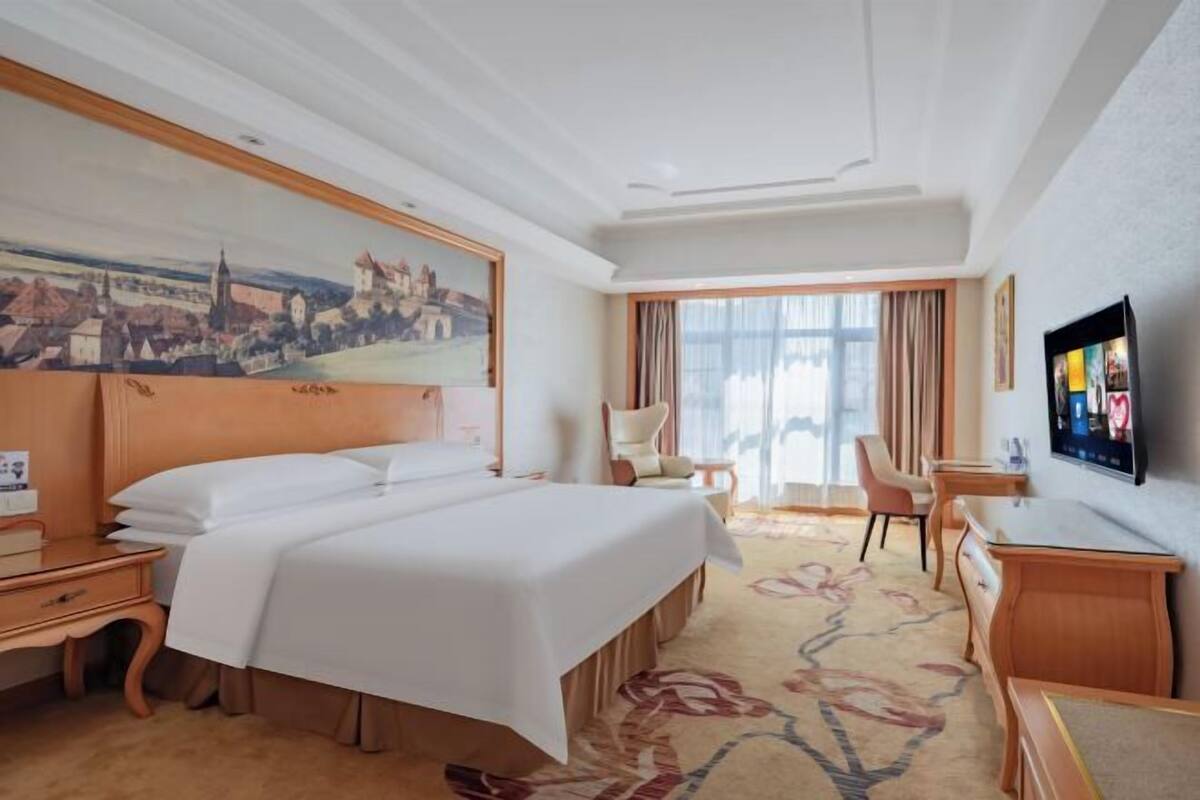 A hotel room with a large bed, a desk, a chair, a TV, and a painting on the wall.