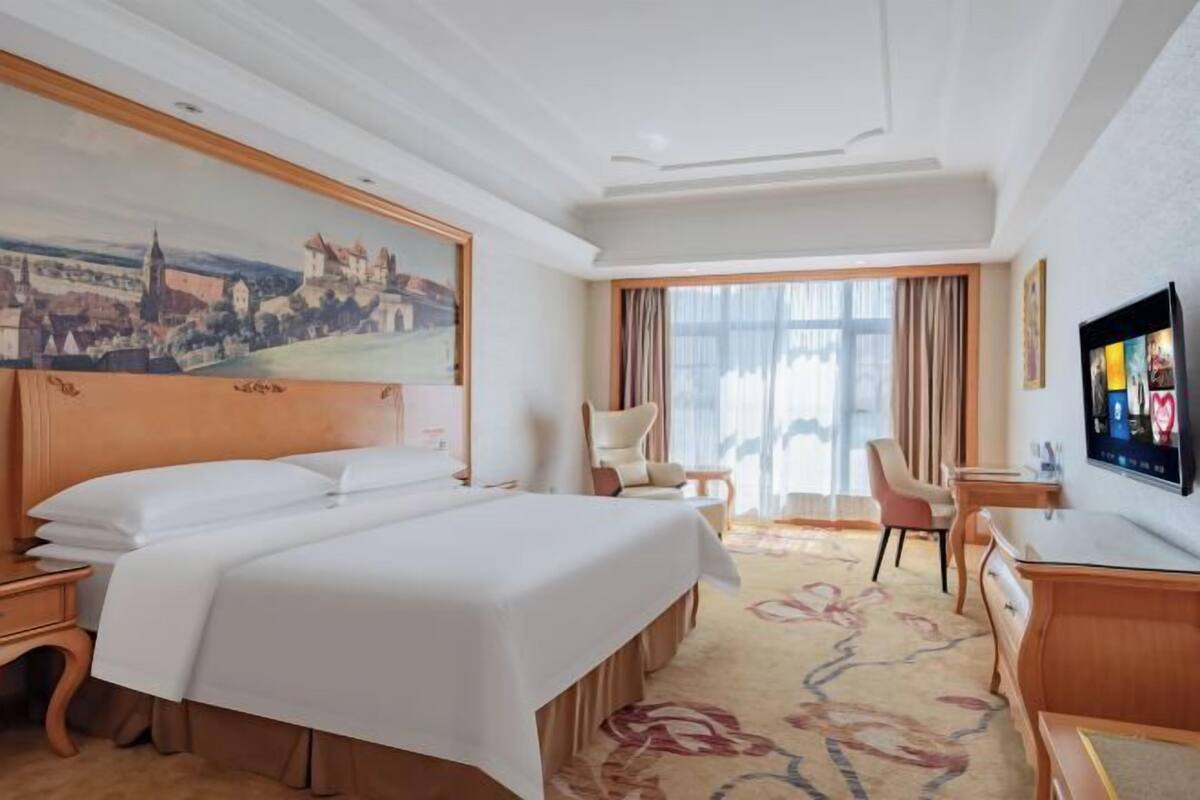 A hotel room with a large bed, a desk, a chair, a television, and a painting on the wall.