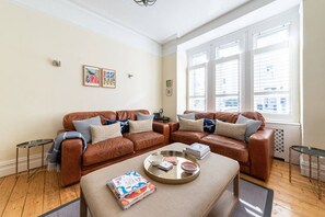 A living room with a brown leather sofa set, a glass coffee table with books and a tray, and a large window with blinds. - Elegant 2-bed Fulham house with private garden (London)