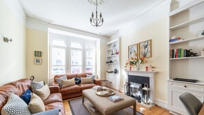 Ein Wohnzimmer mit braunem Ledersofa, einem Holztisch und einem Kamin. Es gibt Bücherregale und eine Pendelleuchte.