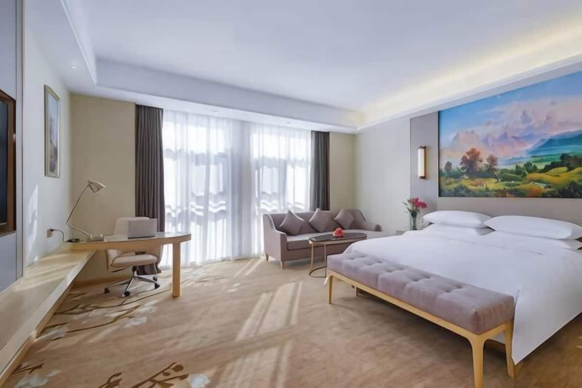 A hotel room with a bed, a desk, a chair, a sofa, and a painting on the wall.
