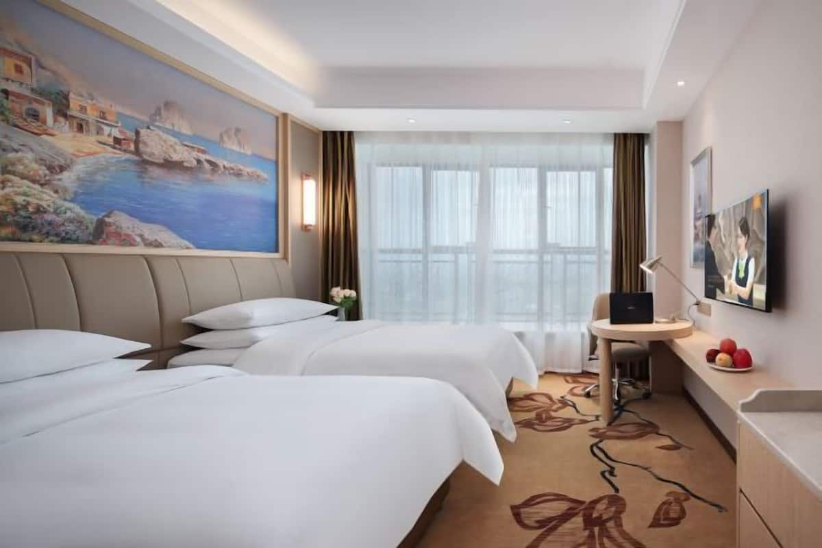 A hotel room with two beds, a desk, a chair, a small table with fruit, and a painting on the wall.