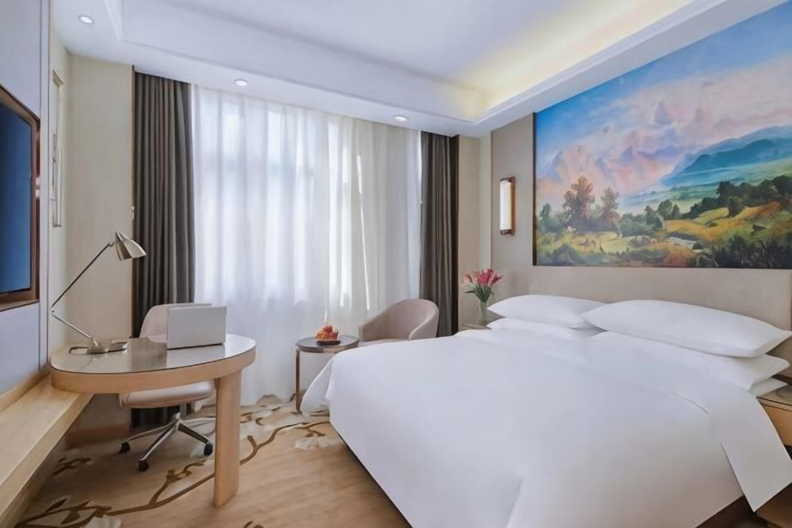 A hotel room with a bed, desk, chair, and a painting on the wall.