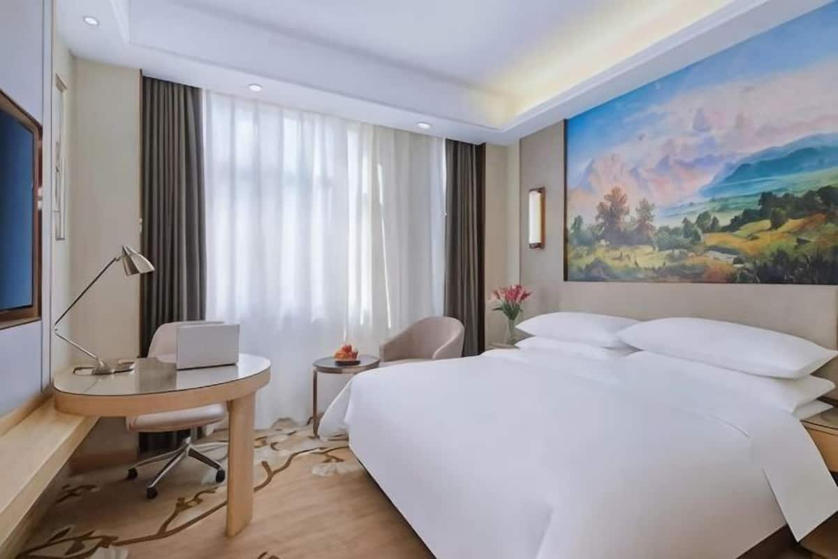 A hotel room with a bed, desk, chair, and a painting on the wall.