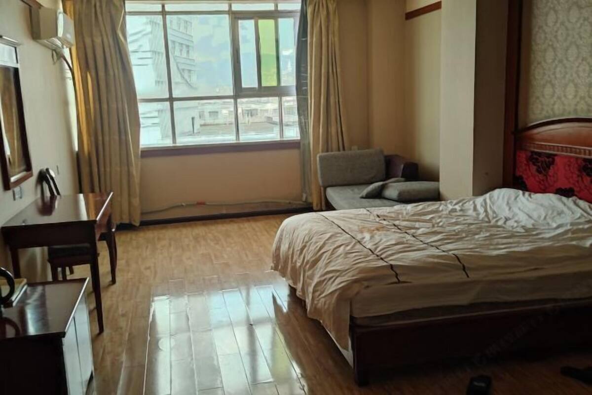 Une chambre d’hôtel avec un plancher en bois, un lit recouvert d’une courtepointe blanche, un bureau avec une chaise et une fenêtre avec des rideaux.