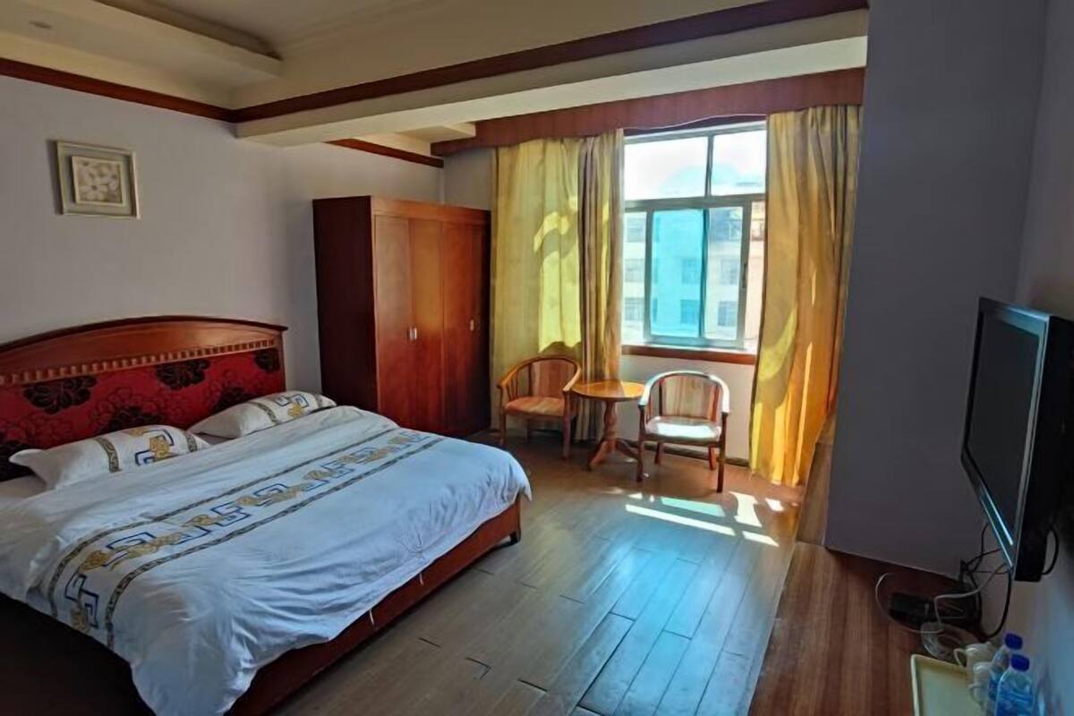 Une chambre d’hôtel avec un grand lit, une tête de lit en bois, une petite table avec des chaises, un téléviseur et une fenêtre avec des rideaux.