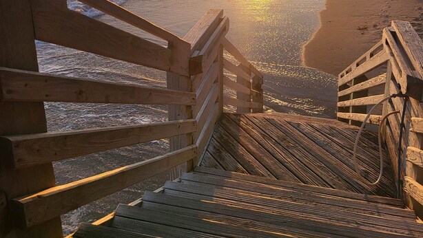 Een houten trap die naar een strand leidt, met een ondergaande zon op de achtergrond.