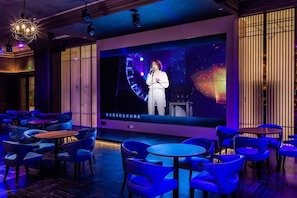 A stage with a performer, surrounded by tables and chairs, in a room with a large screen displaying a cityscape. - Fuqing Yonghong Hotspring Resort (Fuqing)