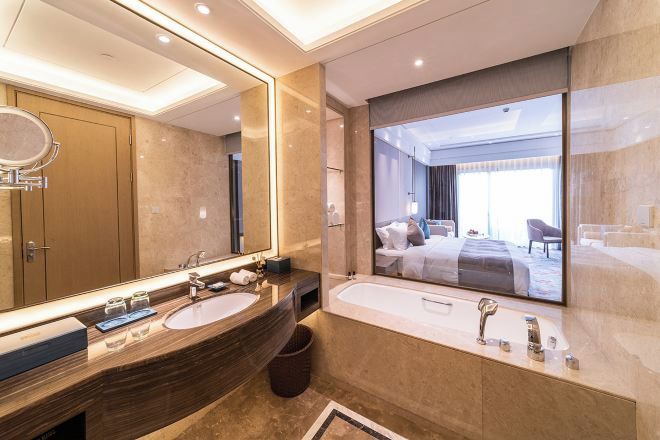 A modern bathroom with a large mirror, two sinks, a bathtub, and a view of a bedroom.