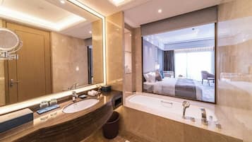 A modern bathroom with a large mirror, two sinks, a bathtub, and a view of a bedroom.