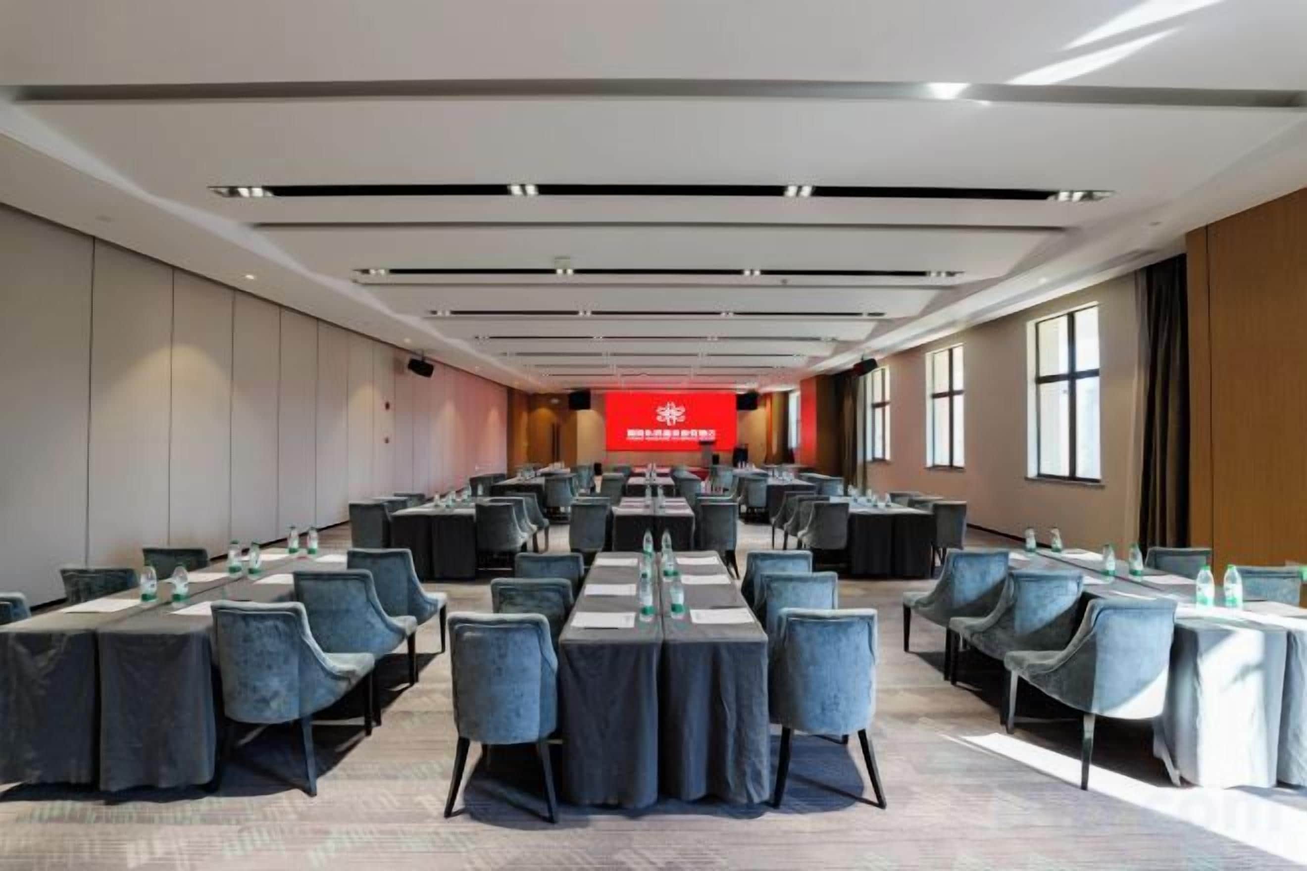 Un salón de conferencias con mesas y sillas dispuestas para reuniones, un banderín rojo con texto, y ventanas amplias a lo largo de una pared.