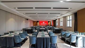 Un salón de conferencias con mesas y sillas dispuestas para reuniones, un banderín rojo con texto, y ventanas amplias a lo largo de una pared.