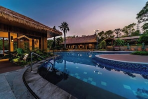 Una piscina con borde curvo, rodeada de una zona de estar con asientos y un edificio con techo de paja.