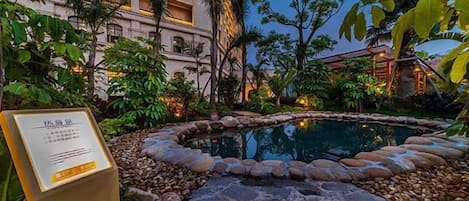 A hotel pool area with a stone border, surrounded by tropical plants and a building with multiple balconies.