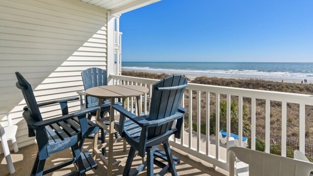 Un balcon abrité, doté de deux fauteuils Adirondack et d’une petite table, offrant une vue sur une plage, l’océan et un ciel dégagé.