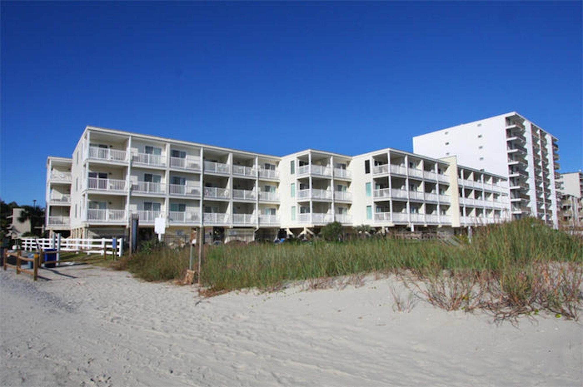 A beachfront hotel with multiple balconies, a sandy beach in the foreground, and a clear blue sky.