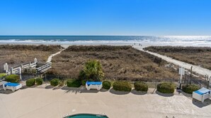Une vue sur la plage avec un chemin en béton menant à une zone de piscine, entourée d’arbustes et d’une clôture.