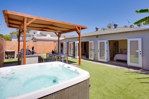 Un jardin avec un jacuzzi, une pergola en bois, une terrasse avec des meubles d’extérieur et une maison dotée de portes-fenêtres coulissantes.