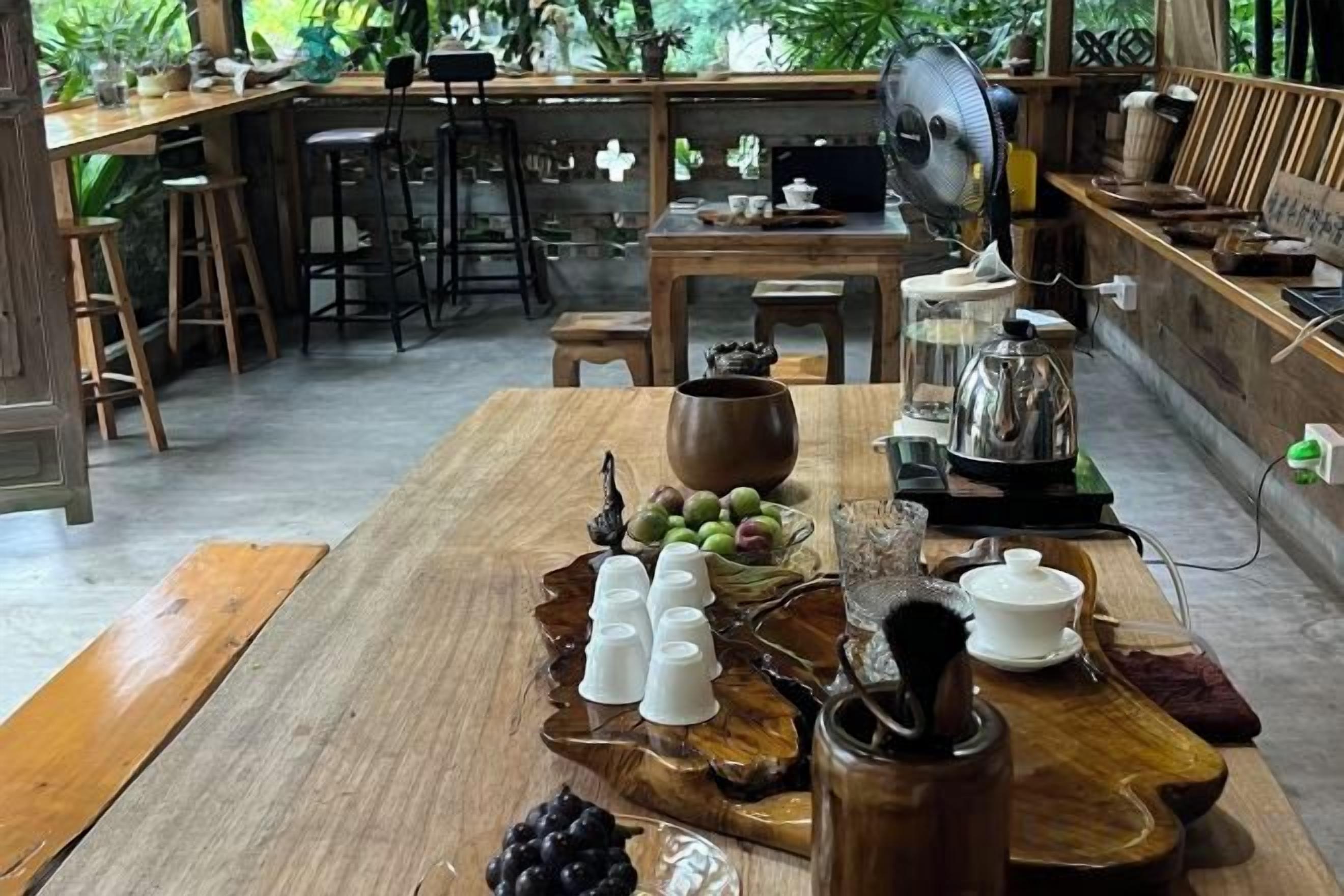 A wooden dining table with a fan, a coffee pot, and a tray with cups and fruits.
