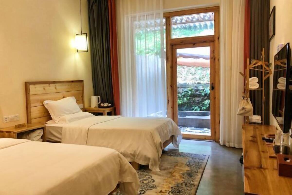 A hotel room with two beds, a wooden headboard, a nightstand, a wooden desk, and a view through a glass door to an outdoor area.