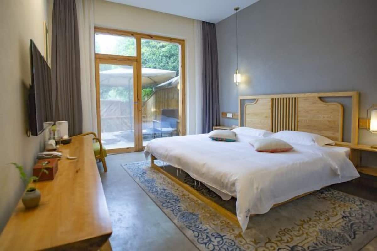 A bedroom with a bed, a wooden desk, a patterned rug, and a view of greenery through a sliding door.