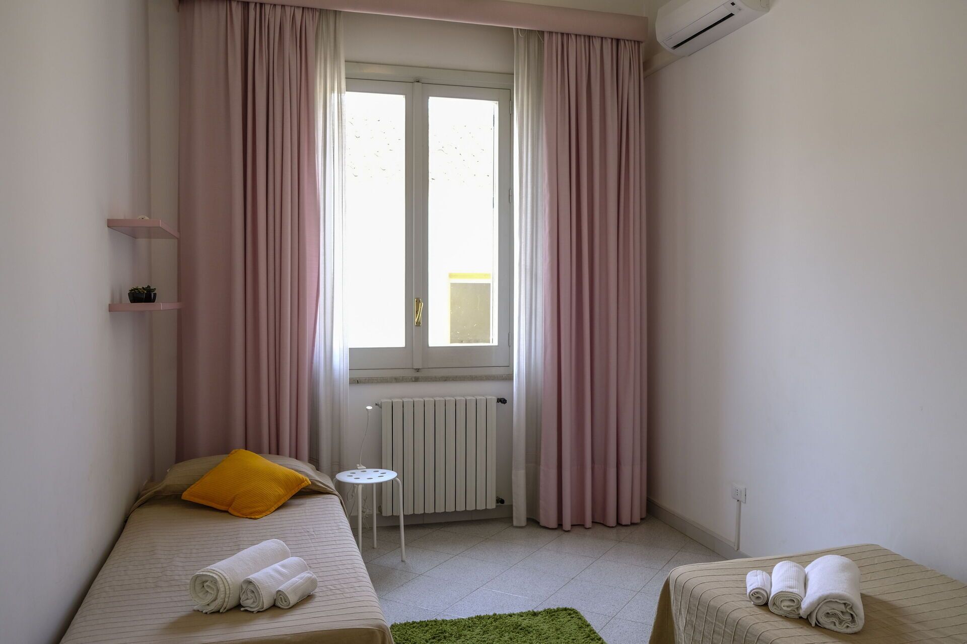 Una habitación pequeña y bien iluminada con dos camas, un radiador, una ventana con cortinas y una mesita.